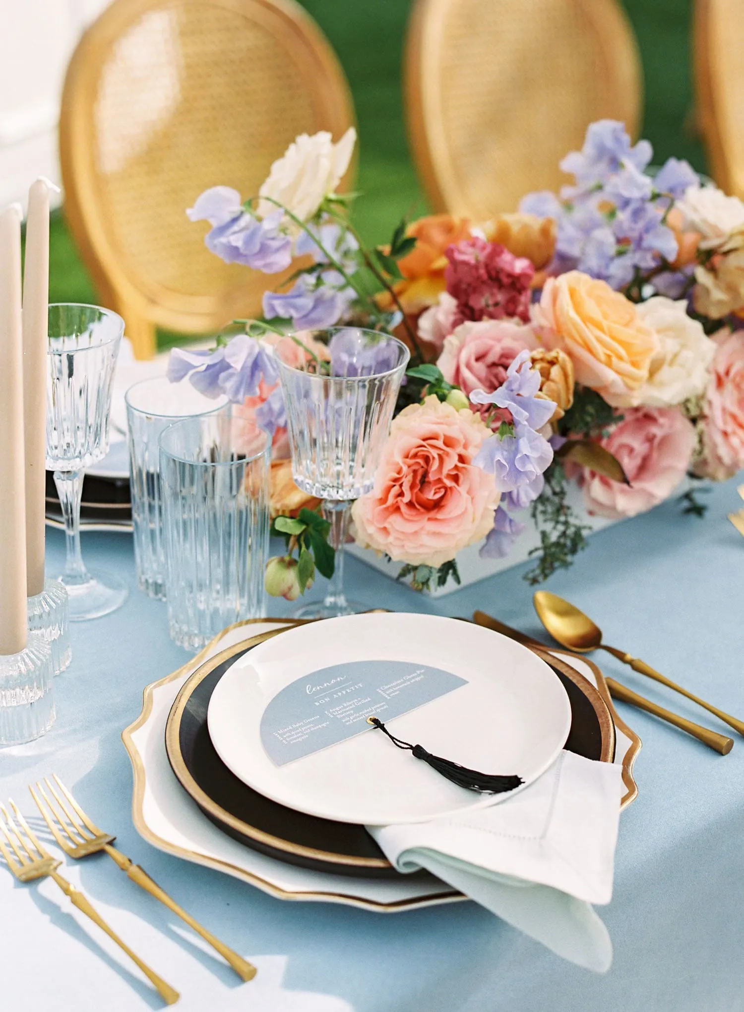 Layered black-and-white china with gold flatware, pale blue linen, crystal glassware, and colorful pastel floral arrangements.