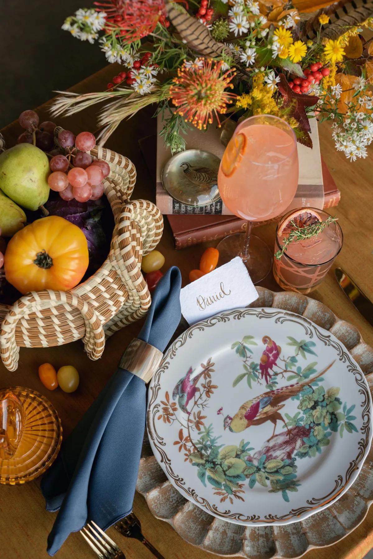 Fall place setting with a bird-patterned plate, navy napkin, seasonal fruit, orange drinks, and colorful autumn flowers.