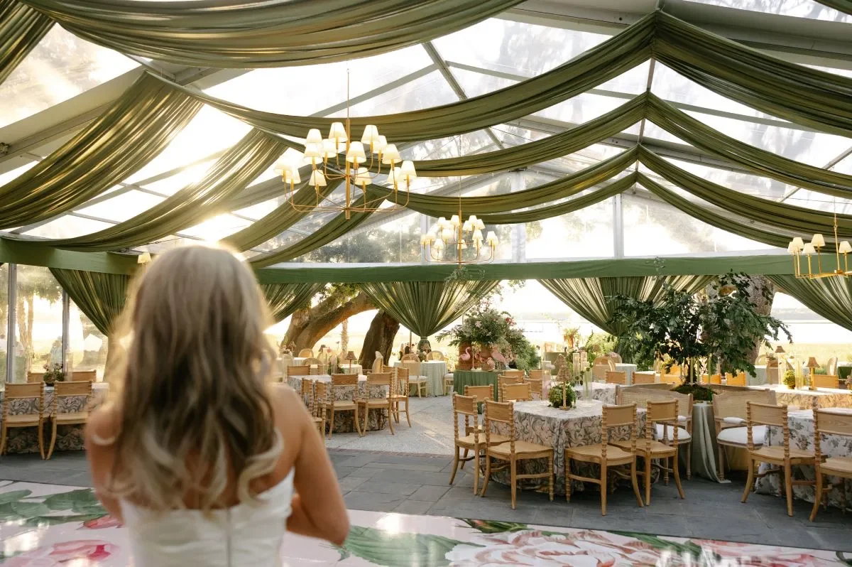 White lounge furniture beneath a clear tent with green draping and brass chandeliers