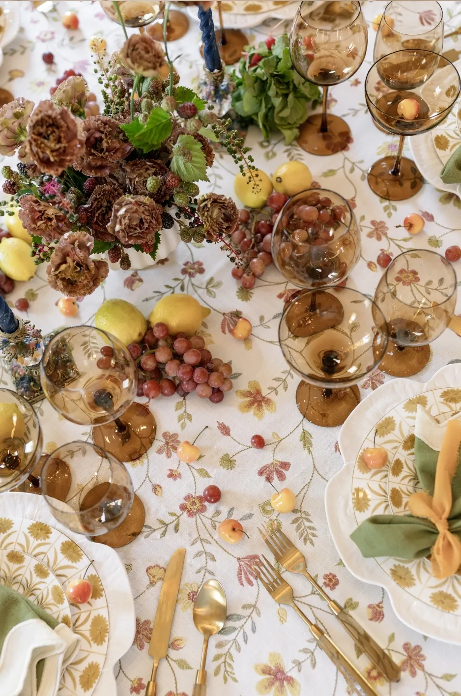 Floral-patterned fall linen with brown flowers, amber glassware, gold flatware, green napkins, pears, grapes, and seasonal fruit.