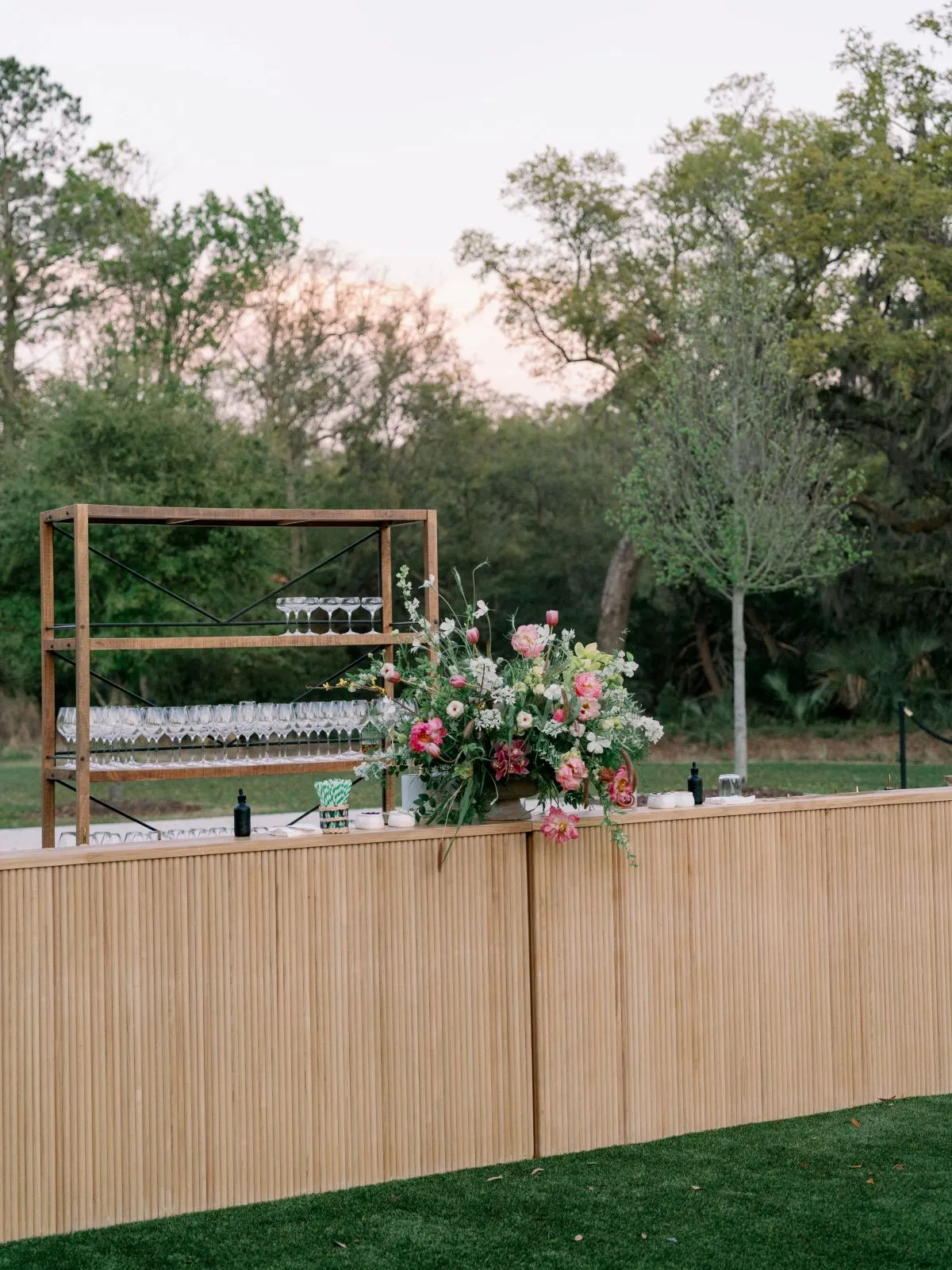 Light wood outdoor bar with a colorful floral arrangement and glassware set against a tree-lined lawn.