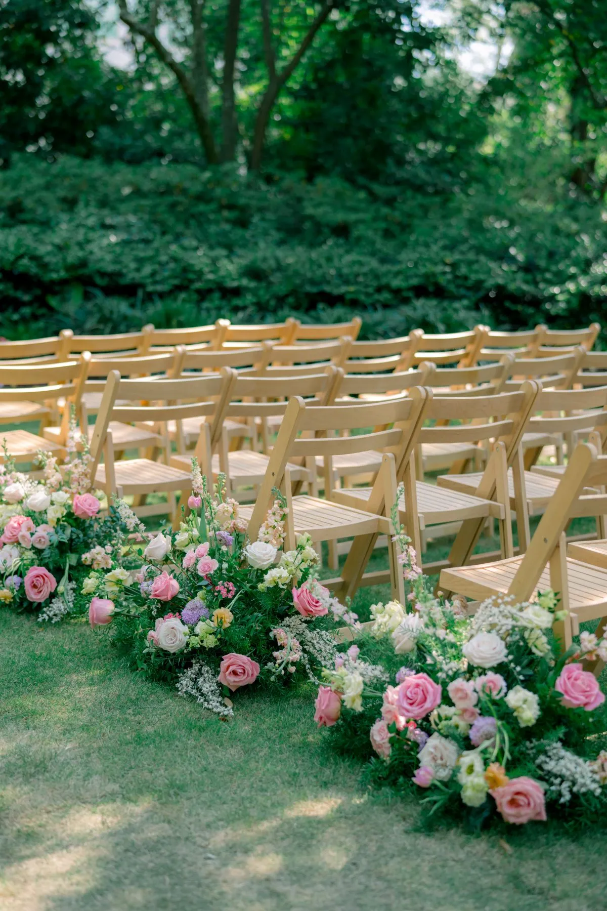 Rows of light wood ceremony chairs beside a low aisle arrangement of pink and white flowers in a garden. | Curated Events Rows of light wood ceremony chairs beside a low aisle arrangement of pink and white flowers in a garden.