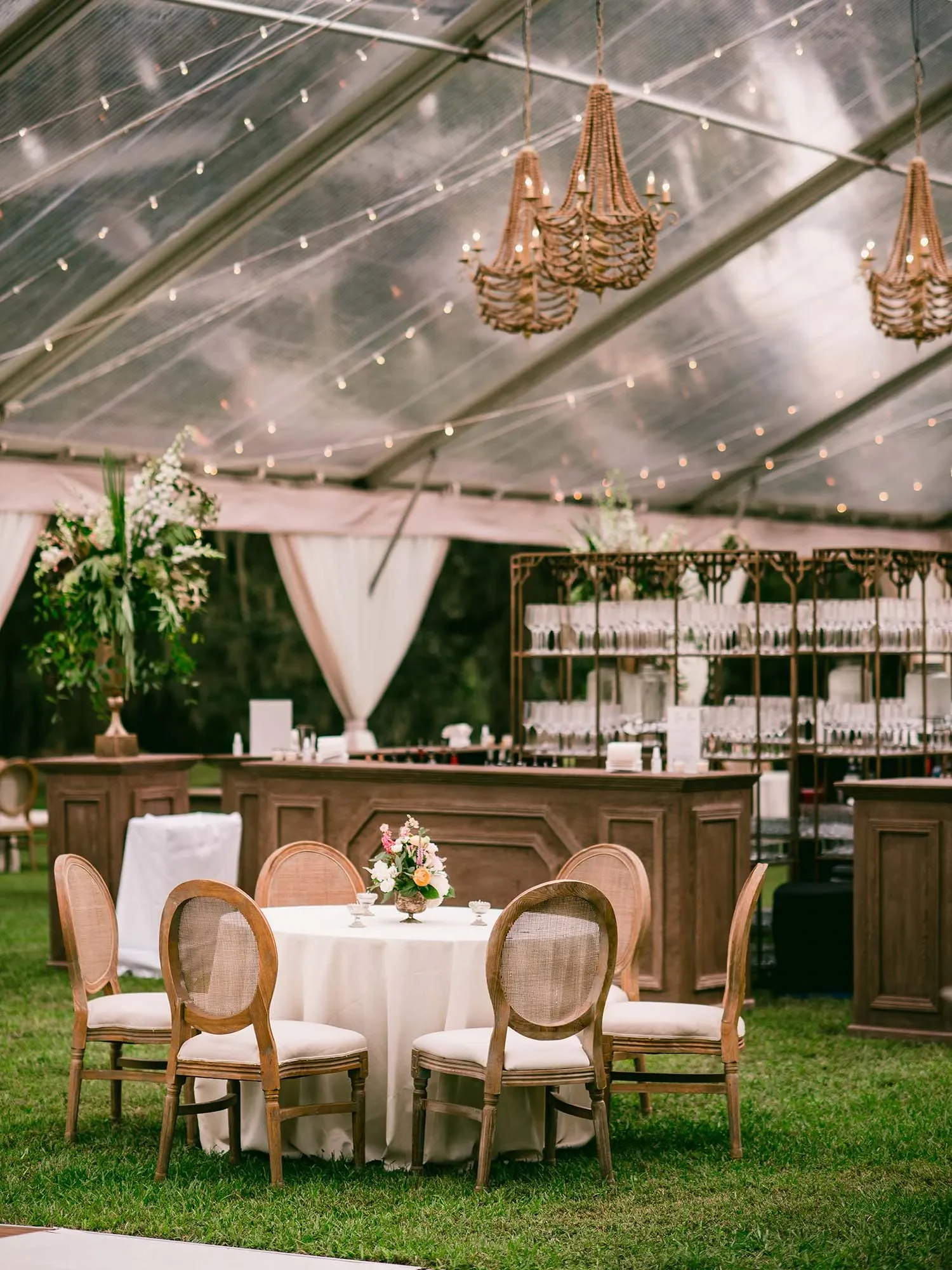 Wood reception bar with backbar shelving beneath chandeliers and string lights inside a clear tent
