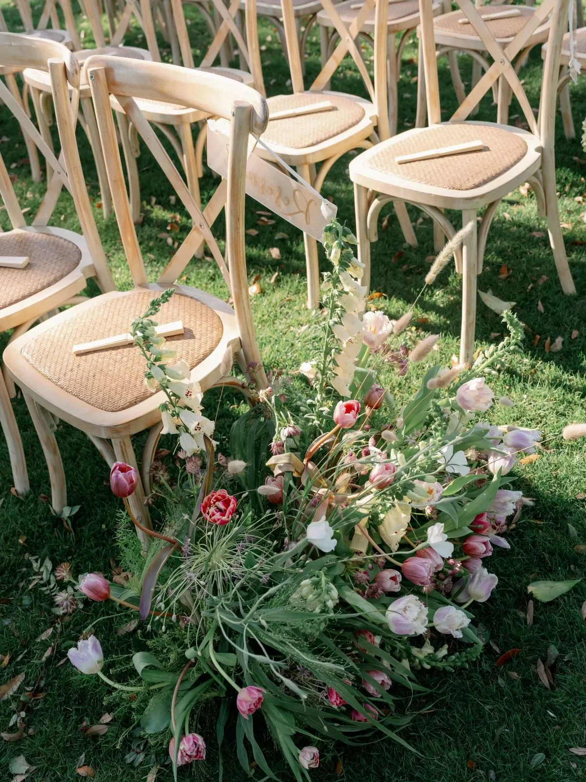 Wooden ceremony chairs beside a colorful ground floral arrangement on a lawn