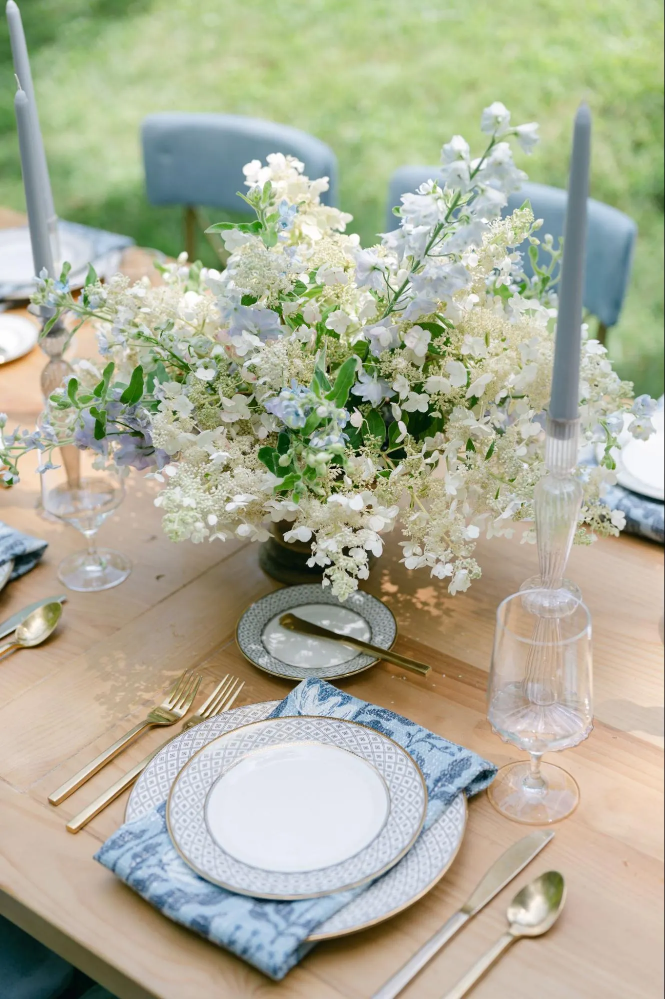 Wood reception table with blue patterned china, ribbed clear goblets, grey taper candles, and white flowers.