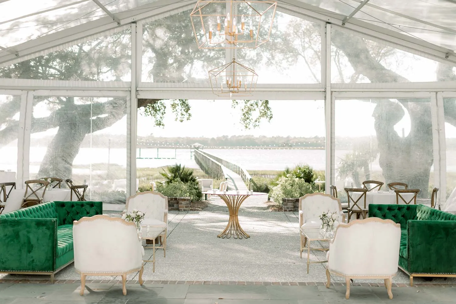 Clear tent lounge with emerald green sofas, cream chairs, geometric chandeliers, and a waterfront view.