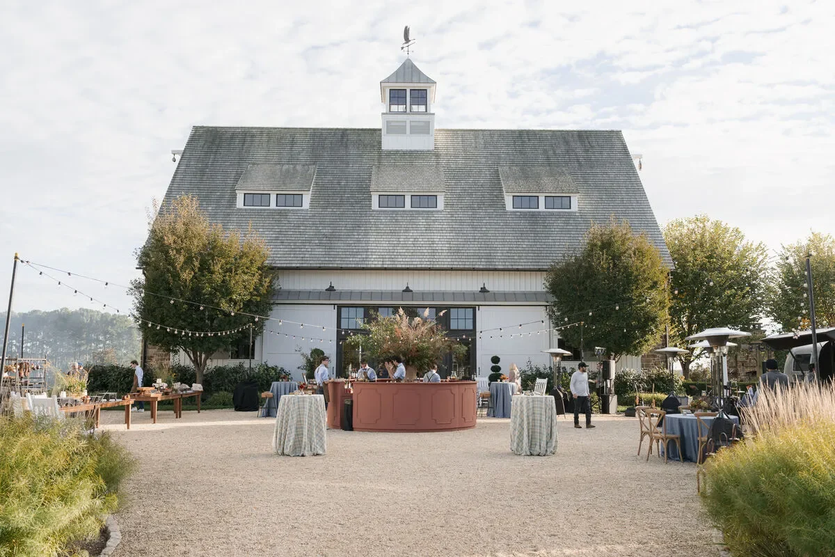 Outdoor wedding cocktail area with a statement bar, cocktail tables, and string lights beside a barn