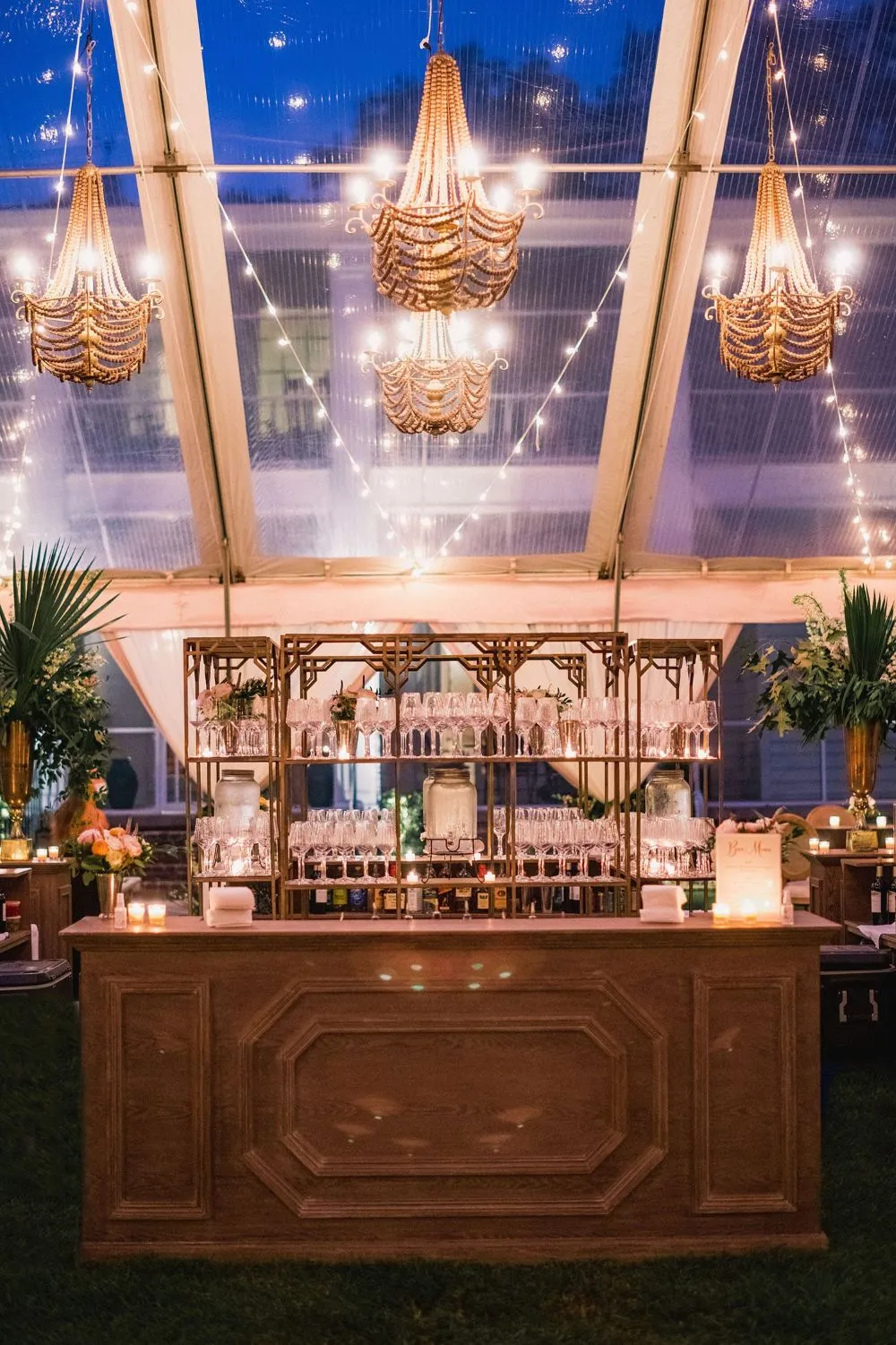 Elegant bar setup under a tent with chandeliers and string lights, surrounded by floral arrangements